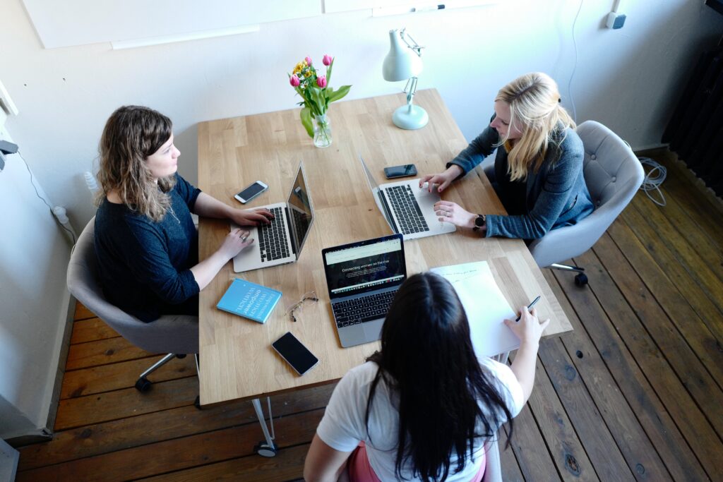 In this image, three businesswomen are shown seated around a table, engaged in a discussion. All three women are dressed in professional attire, and each has a laptop open in front of them. The laptops are positioned on the table, and the women are leaning in towards one another, suggesting that they are closely collaborating on a project. The women have serious expressions on their faces, indicating that they are focused and dedicated to their work. The image conveys a sense of teamwork and professionalism, as well as the importance of technology in modern business.